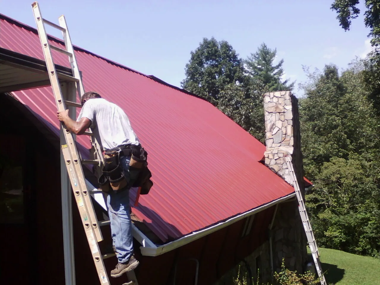 Roofer on a ladder beside a red metal roof and stone chimney — Soffit & Fascia Repair by Roofing Brothers in Pilot Rock